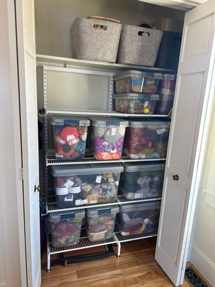 Photo of my yarn closet after a big purge. It's a small bedroom closet with bifold doors, one built in shelf up top, and a white wire shelving unit with four shelves sitting inside. On the wire shelves are plastic bins full of yarn. On the built in shelf are felt bins full of photo props. It is uncharacteristically tidy, this state will not last.