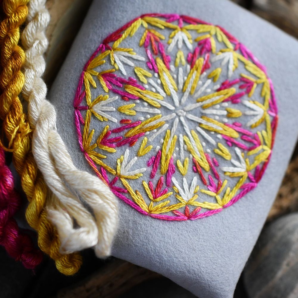 Photo of embroidery. The embroidery is on gray felt using pink, yellow, and white silk embroidery thread. The design is a circle with the same design repeated on eight points to make sort of a snowflake/kaleidoscope/round stained glass window sort of vibe.