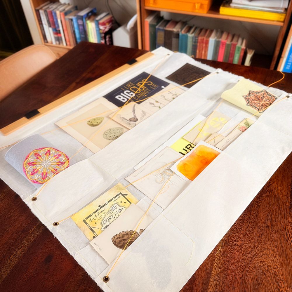 Photo of a wall hanging made from one giant sheet of white paper, folded up into several rows of pockets, with various bits of pretty papers tucked in the pockets. It's laying on my dining room table and you can see my bookcases in the background. The other pictures in the thread are more pictures of the same thing from other angles. 