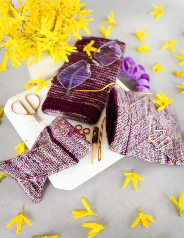 photo of three knitted pouches of different sizes and designs, arranged on a gray table, with various small knitting notions scattered about. there are also some pretty yellow flowers for good spring vibes.