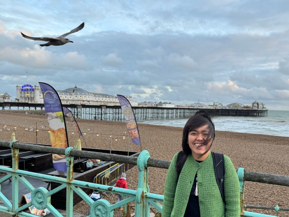 Asian woman in green jacket standing in front of the beach and a seagull approaching