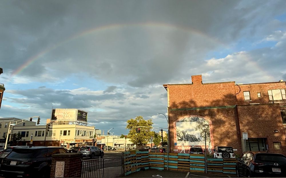 A wider view of the full rainbow, which comes down somewhere behind a brick building with a mural on the side. 