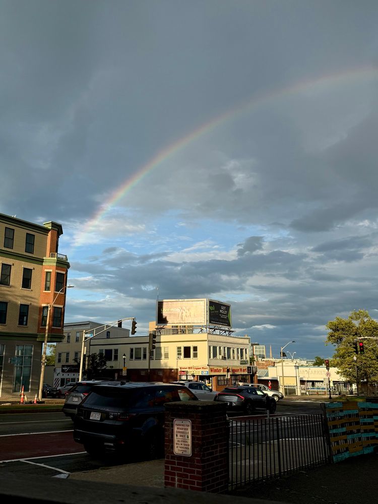 The left half of a rainbow arcs over a city street. A few bright blue patches of sky peek out between gray clouds. 