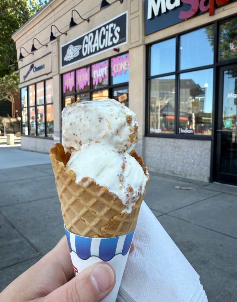 A white hand holds a waffle cone with two scoops of white ice cream that has brown and black bits in it. In the background is the storefront of Gracie’s ice cream. 