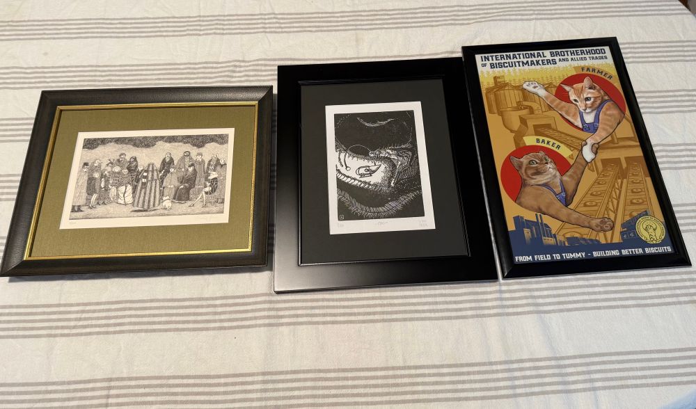Three pieces of framed art on a table. On the left, a line drawing of a group of people. In the middle, a linocut print of a deep-sea creature eating a submarine. On the right, a poster of two cats in the style of an old Soviet propaganda poster. 