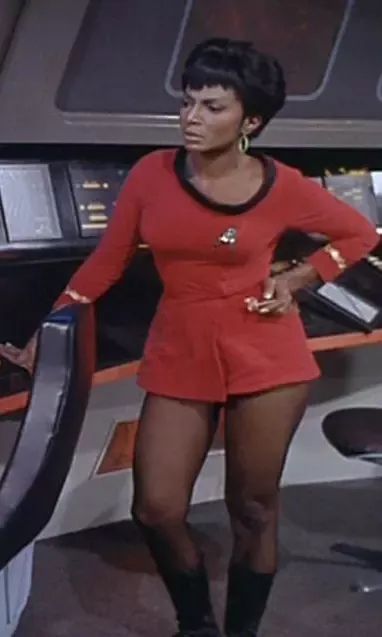 Photograph of lieutenant Uhura from StarTrek: The Original Series.

She is a Black woman in a red dress with 3/4 sleeves and a mini skirt. Under the skirt she wearing low denier tights and calf high boots.