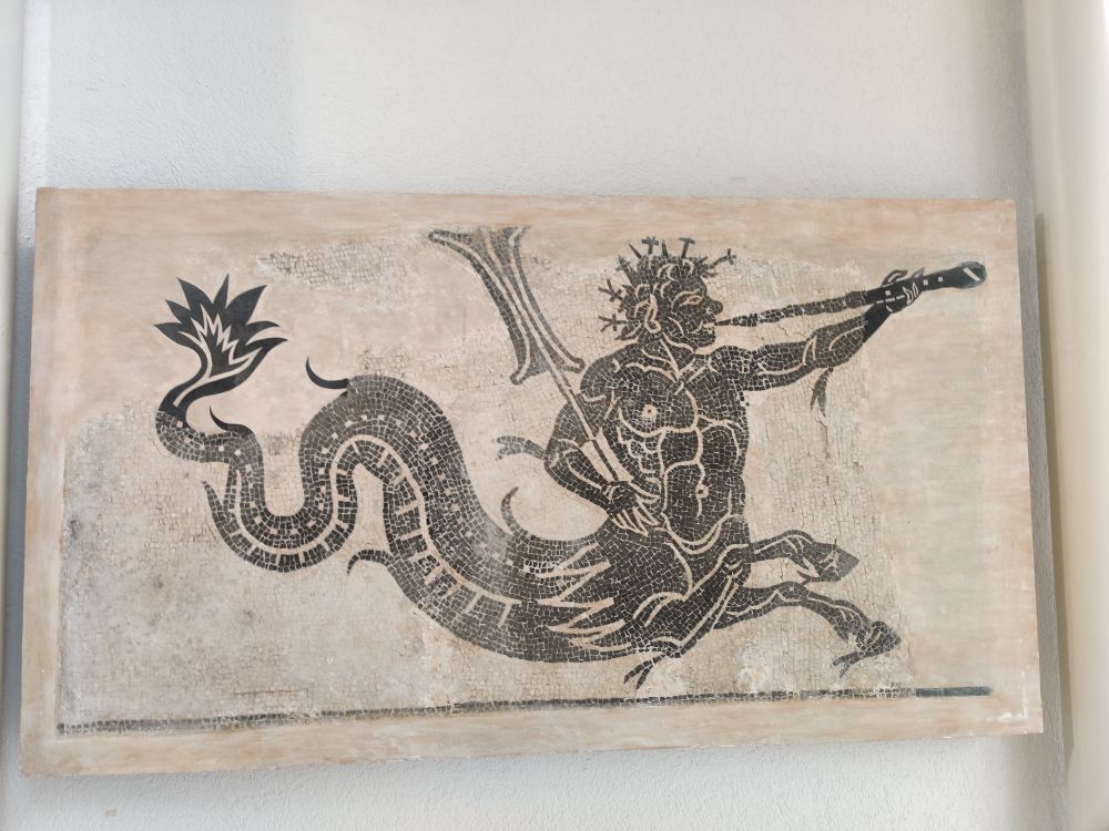 A black and white Roman mosaic depicting a half man - half dragon (or fish) with two front hooves. He carries a pipe or flute type instrument in his left hand and a kinda sign in his right 