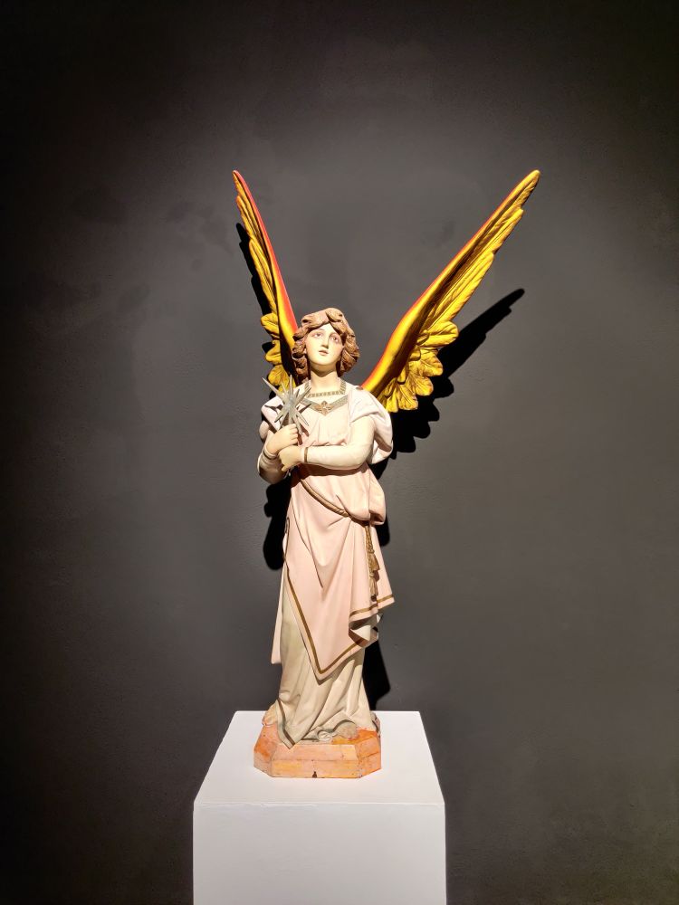 A white statue of a robed angel with yellow wings. It stands on a white pedestal against a black background 