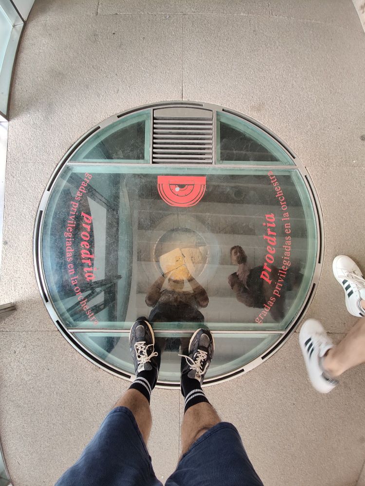 Looking down into a circular, glass, porthole in the Roman theatre visitor centre. My feet and Ronny's feet are visible and our reflections are in the glass. Beyond the reflection you can see a piece of one of the columns that used to support the upper part of the theatre