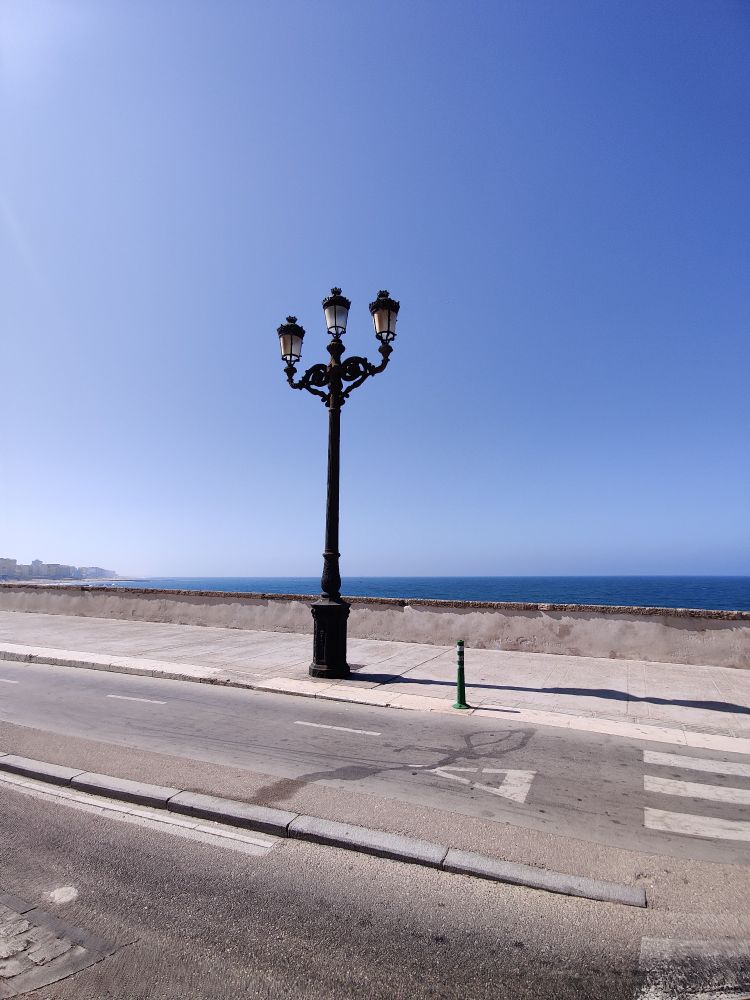 A roadside lamp shaped like a trident, with the sea in background and the coastline road in the fore