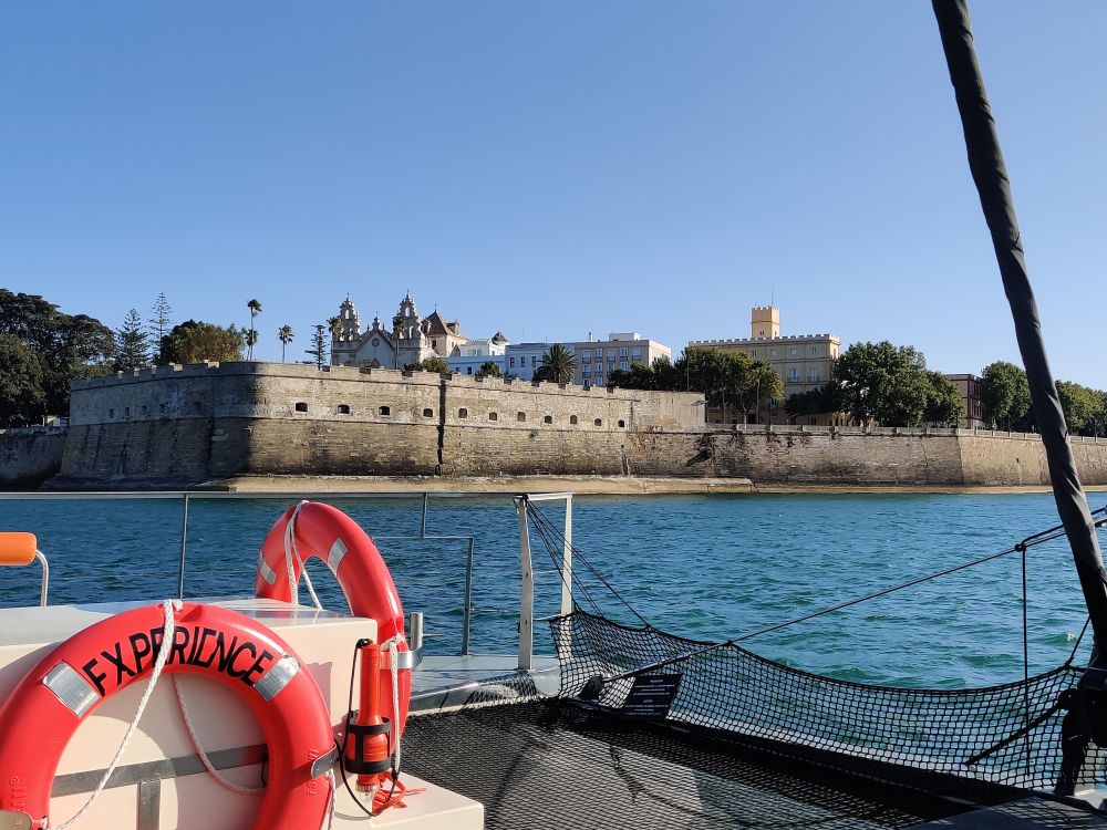 A view of Càdiz city wall  across the front of the catamaran. The wall was built at the coast to protect the city from invaders. There is a row of holes in the wall where cannons could be fired from. In the foreground there are large red life lifebuoys 