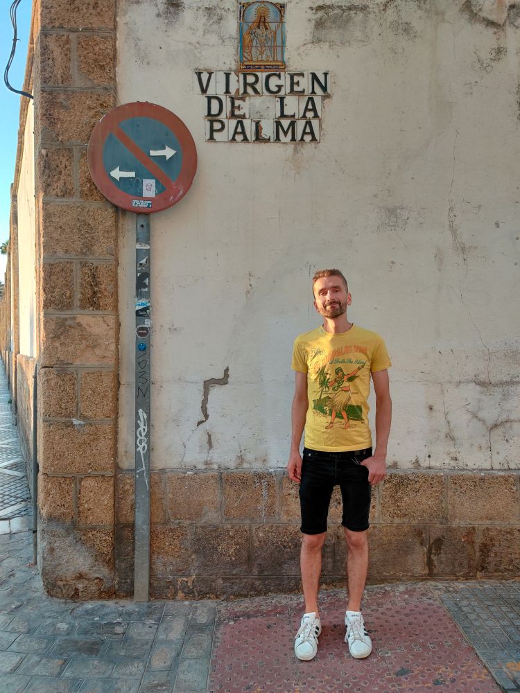 Ronny stands at a street corner under a sign with the street name Virgin de la Palma. This suggests Ronny is the virgin of Palma