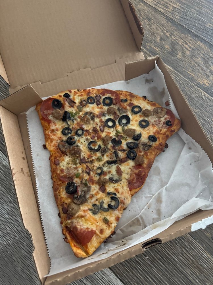 A large slice of pizza in an open cardboard box, resting on a sheet of parchment paper. The pizza is topped with melted cheese, pepperoni, black olives, sausage, and green peppers. The box is placed on a wooden surface.