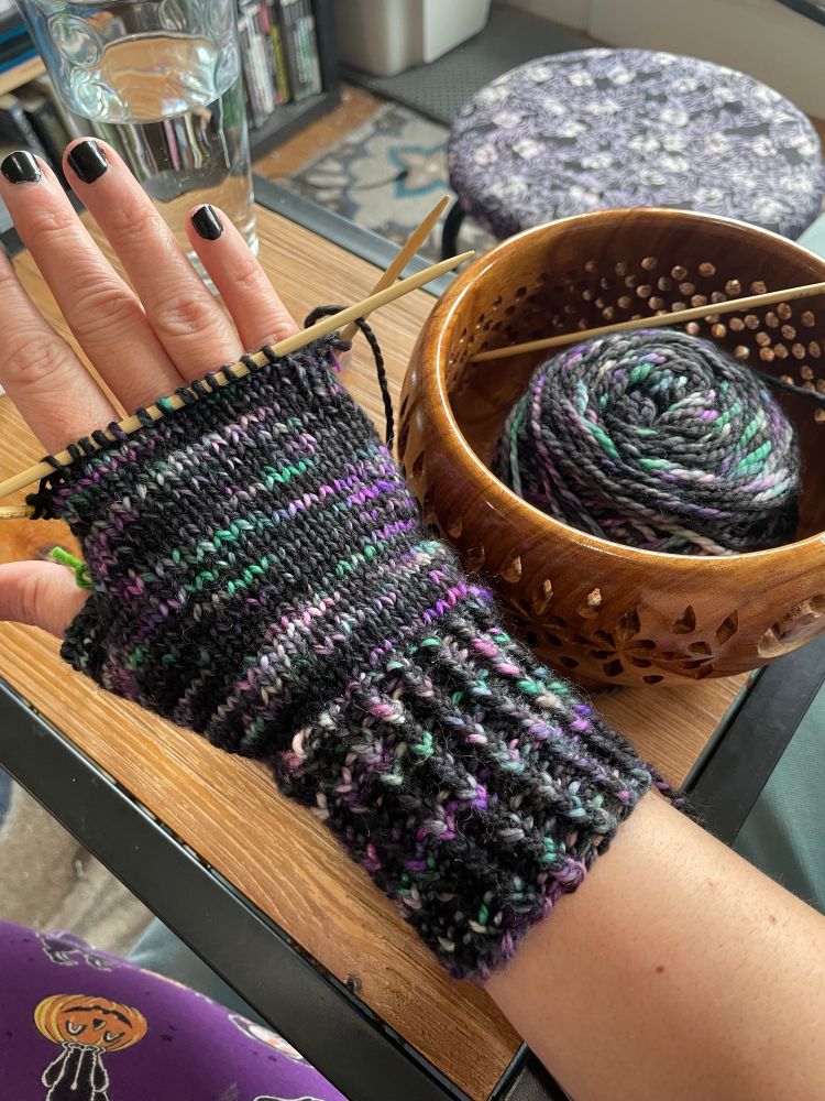 A hand with black nail polish wearing an in process fingerless glove that is still on double pointed knitting needles. To the side is a wooden yarn bowl containing the working yarn: a dk weight variegated yarn that is mostly black with purple, teal, and some white. 