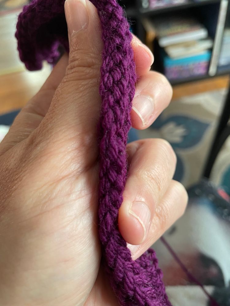 A hand holding purple yarn knit into 12 rows of garter stitch. The person’s middle finger indicates a mistake in the cast on edge. 