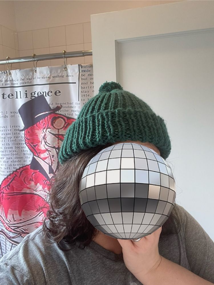 A woman with brown hair is wearing a green k1, p1 rib knit hat. It has a folded brim and a puff ball at the tip. The disco ball emoji is being used to obscure her face. In the background is a shower curtain with a pink T Rex wearing a black top hat. 
