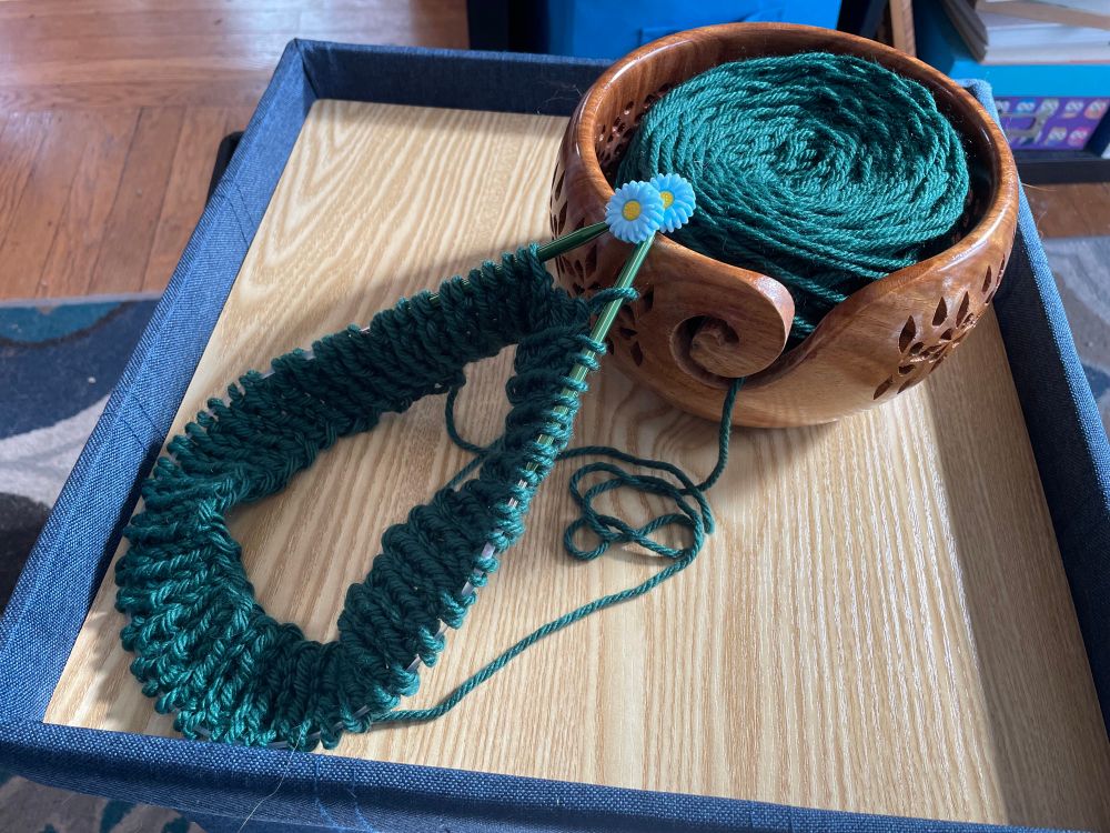 Green chunky yarn in a wooden yarn bowl with the beginning rounds of a simple knit 1, purl 1 hat on circular knitting needles. 