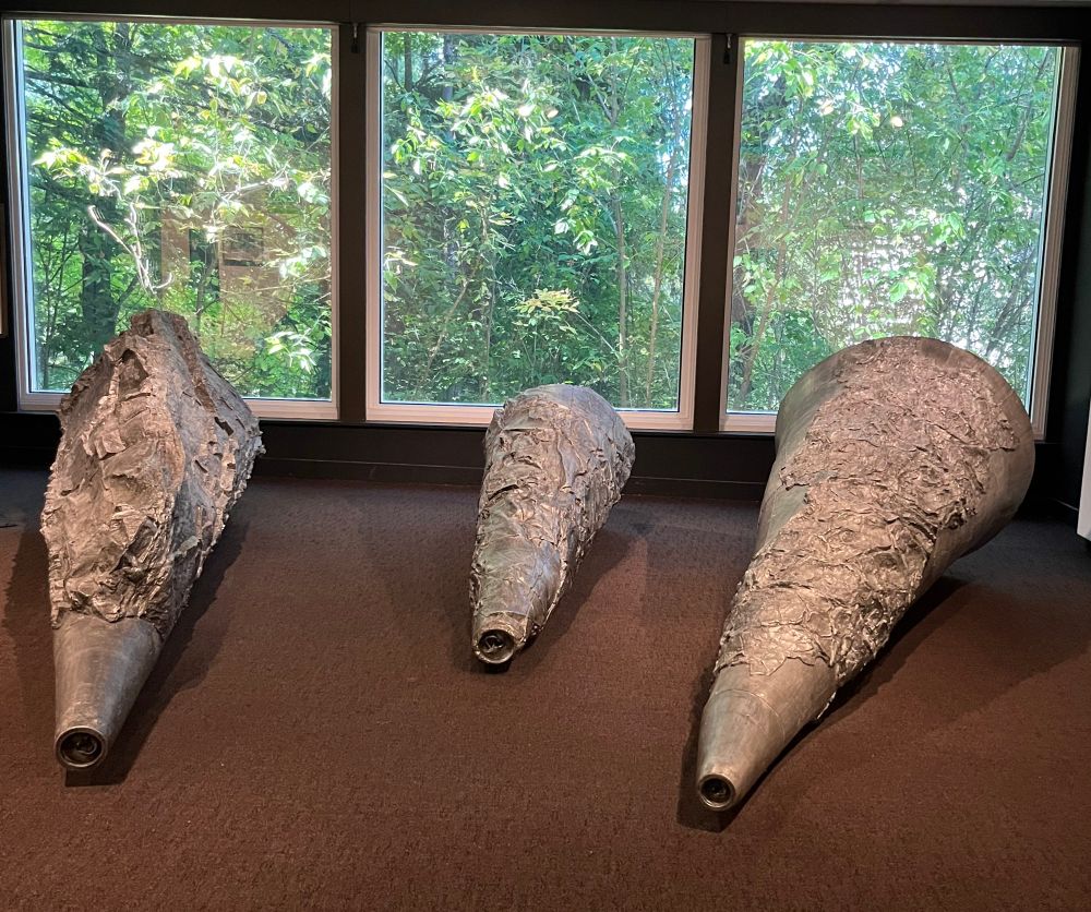 three large cast aluminum megaphone-shaped sculptors sit on floor facing out three windows to forested landscape  