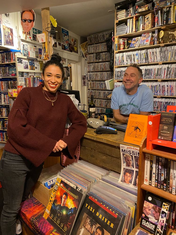 Ellen E Jones standing in front of the counter at 20th Century Flicks in Bristol, UK, the world’s oldest continuously running video shop. Shop owner David Taylor stands behind the counter. Both are smiling broadly.