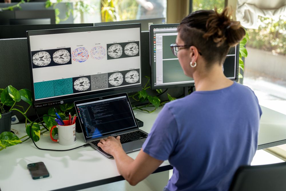 A woman sits at a desk working on a laptop while medical imaging data, such as MRI brain scans, is displayed on a large monitor.