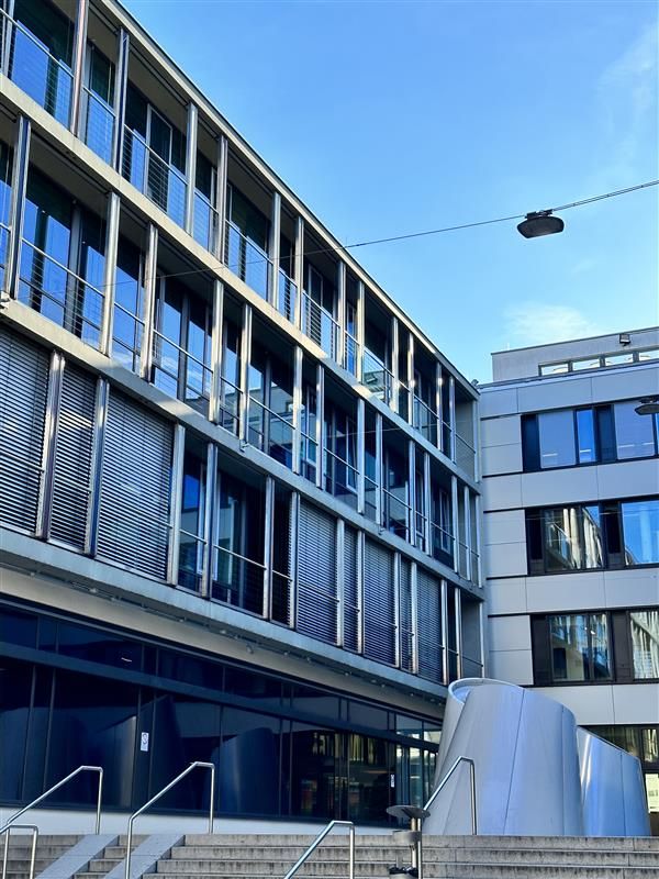 Modern building facade with large glass fronts, slatted blinds, and metallic elements. In the foreground, stairs and a sculptural metal design feature.