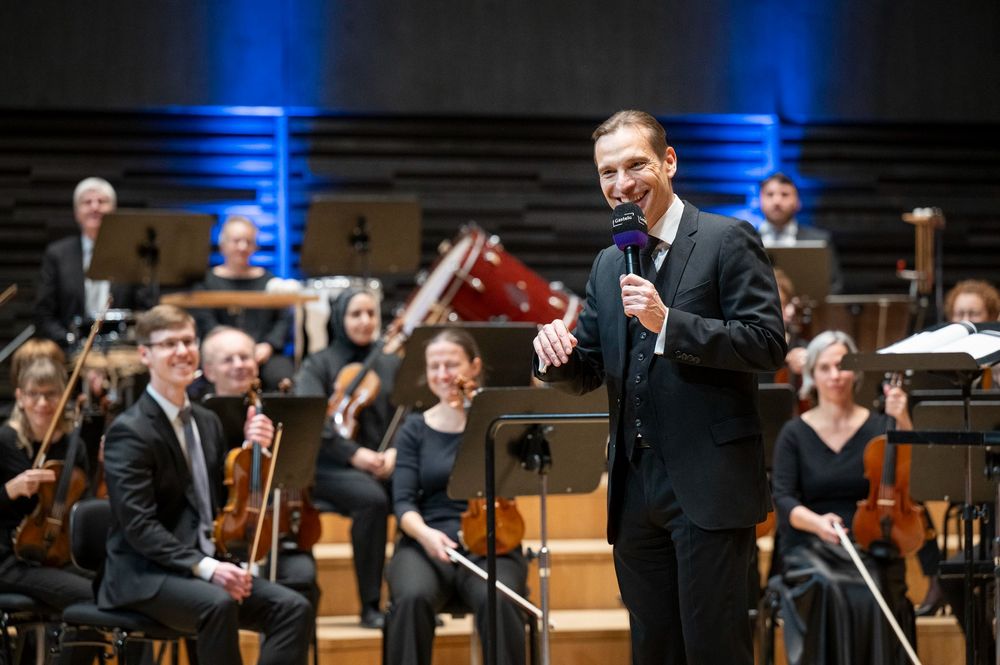 A person in a formal suit speaks into a microphone on stage during the TUM Advent Matinee at Gasteig HP8 / Isarphilharmonie. Behind them, members of an orchestra are seated with string instruments and music stands, under blue stage lighting.