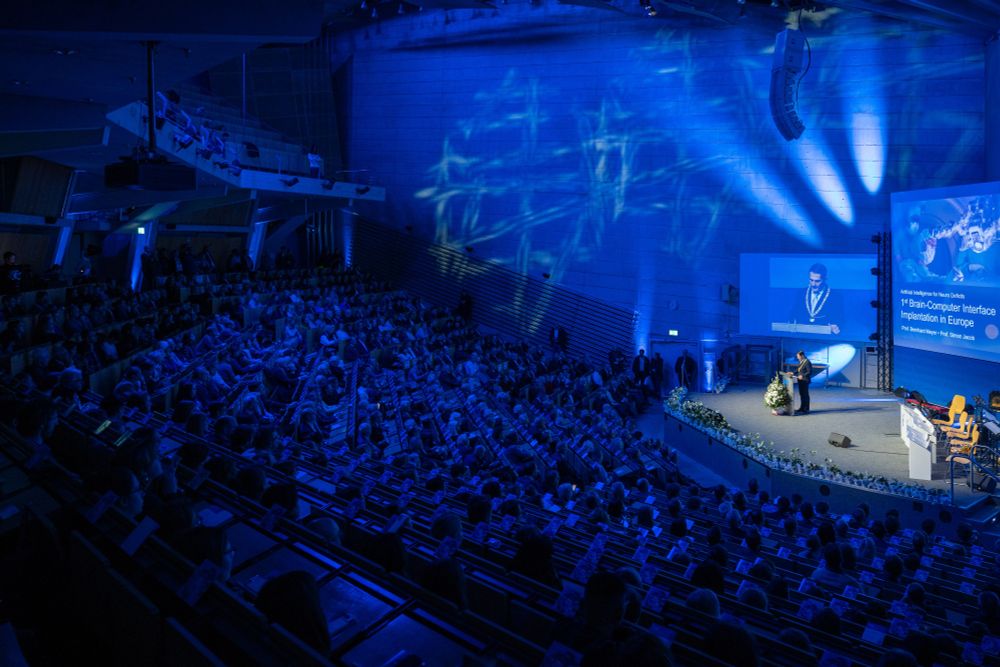 Wide view of the grand lecture hall at TUM during Dies Academicus 2025, illuminated in blue lighting, showing a full audience and a speaker addressing the crowd from the stage.