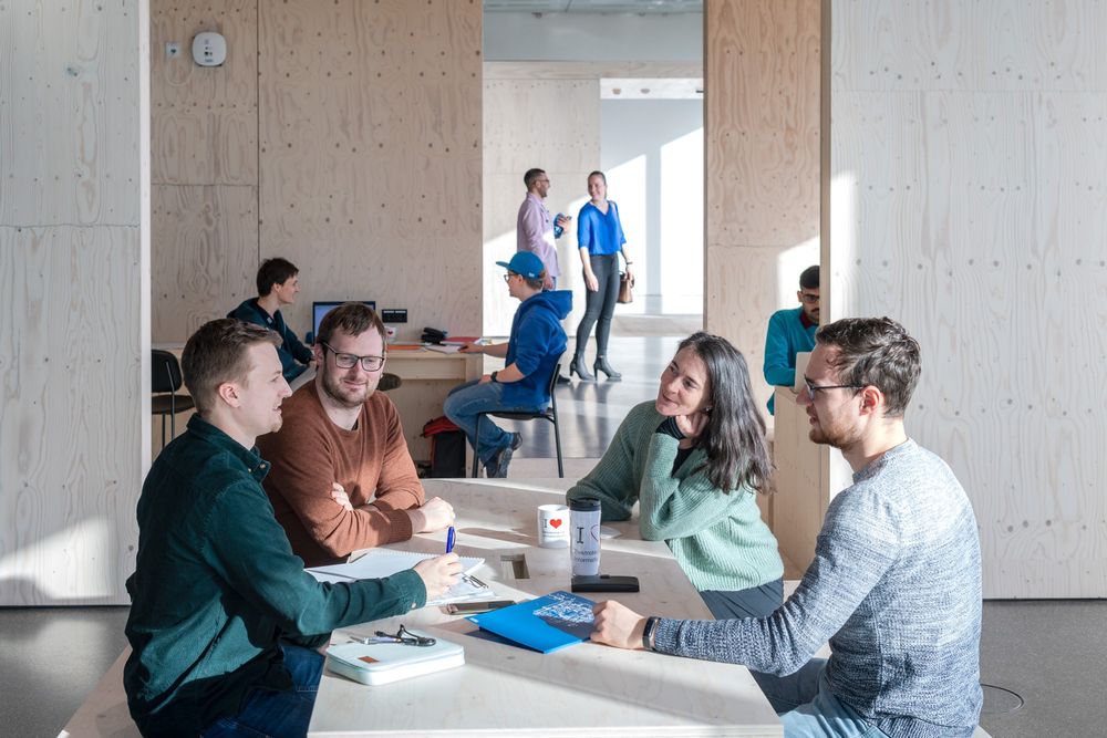 A bright study space with wooden walls, where four people sit around a table, engaged in discussion. In the background, others work or walk through the sunlit room.