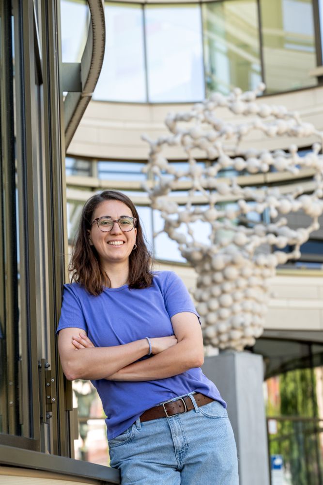 A woman stands smiling with arms crossed against a building facade, with a large sculpture resembling a DNA helix visible in the background.