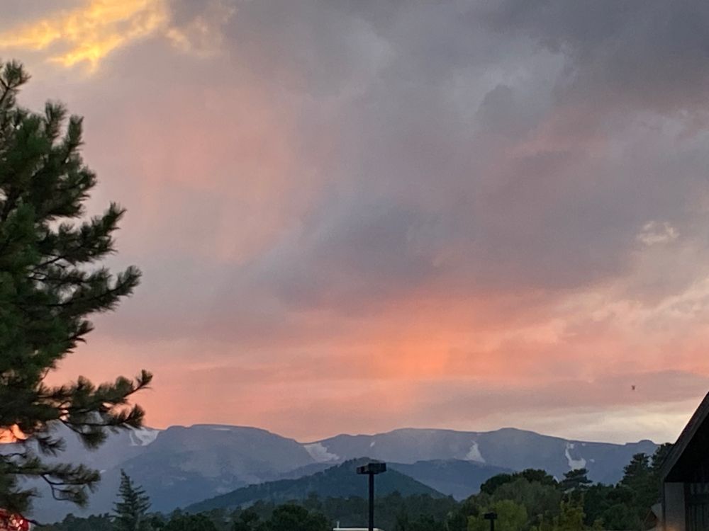 The Rocky Mountains, with cloudy sunset sky above. The blue clouds are painted pink, orange, purple, and yellow by the setting sun and are wispy with drizzle. Toward the left of the frame is a photobombing pine tree.