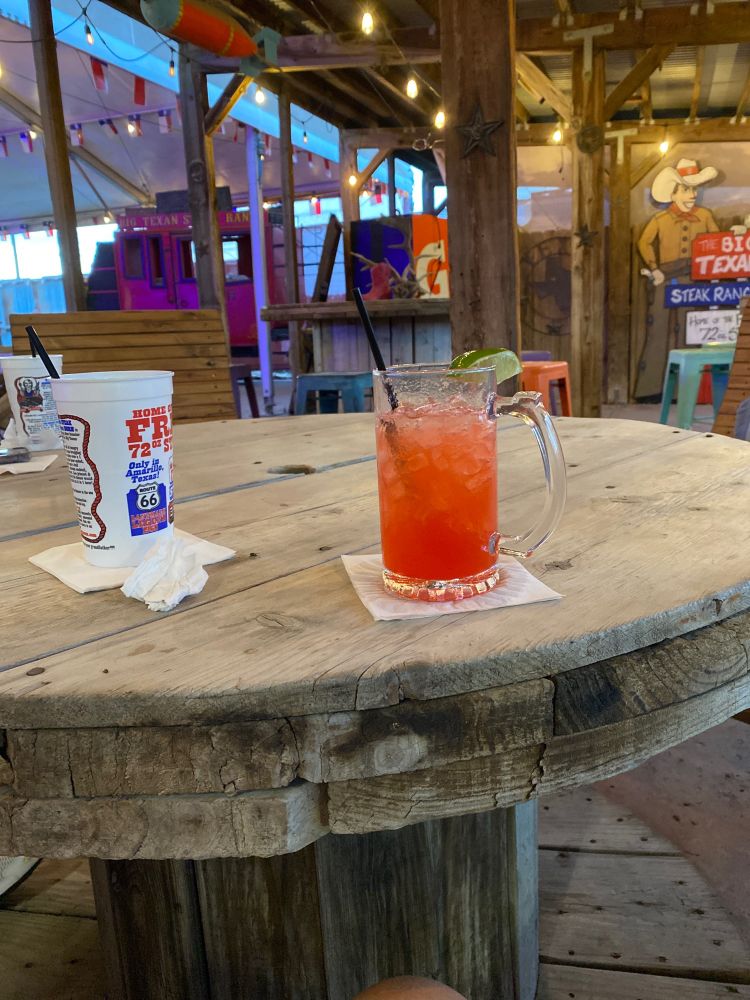 The outdoor space I’m chilling in right now. Photo shows round wooden table with a strawberry something alcoholic drink and my cup of water (gotta hydrate). There’s a roof over us with party lights and other tables in the background.