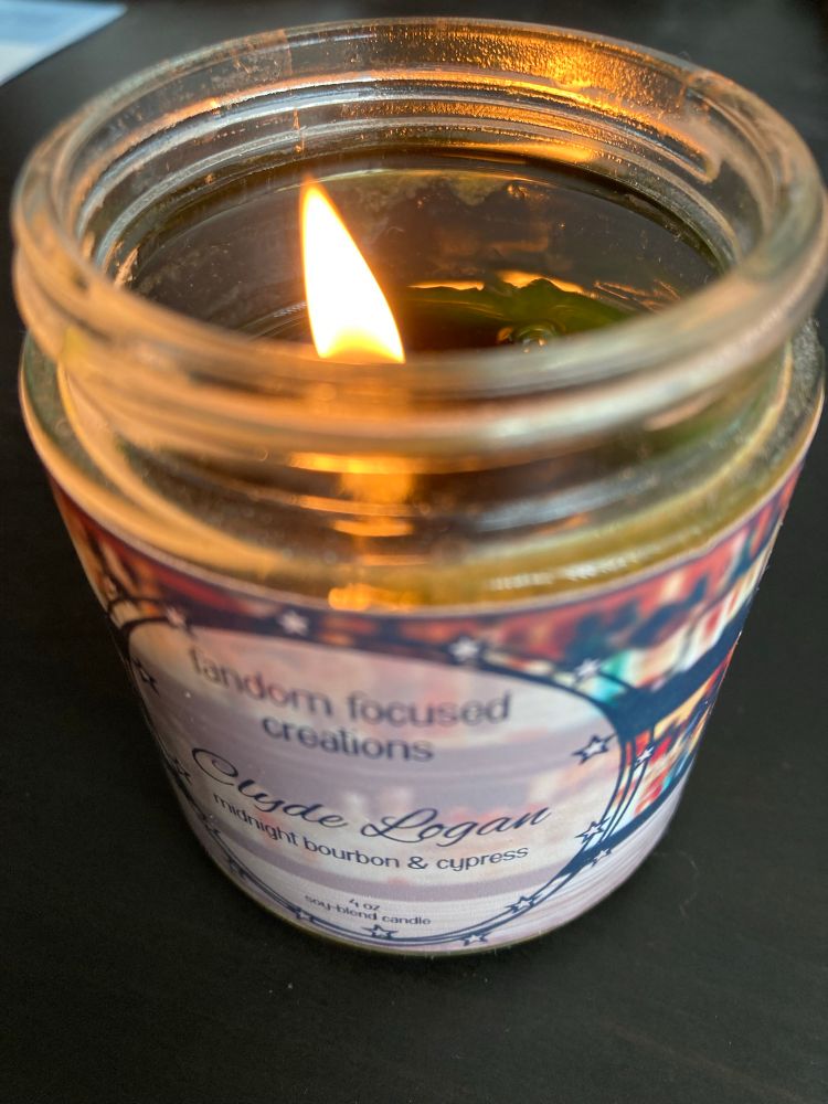 Photo of a candle by “fandom focused creations” called Clyde Logan with scent of midnight bourbon and cypress. 