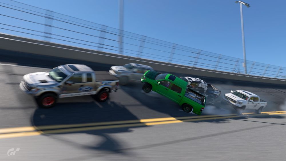 Screenshot from a video game. A green truck has only one wheel on the ground. It it facing 45 degrees away from the track, and colliding with three white pickup trucks. Smoke is emitting from where one corner of the green pickup truck's rear bumper is now rubbing against the asphalt at roughly one hundred miles per hour.  One is thankful that this is a screenshot from a video game and not a real crash involving real vehicles.  It appears that everyone involved just watched their entire day get ruined, their racing trucks get destroyed, and their dreams get shattered. 