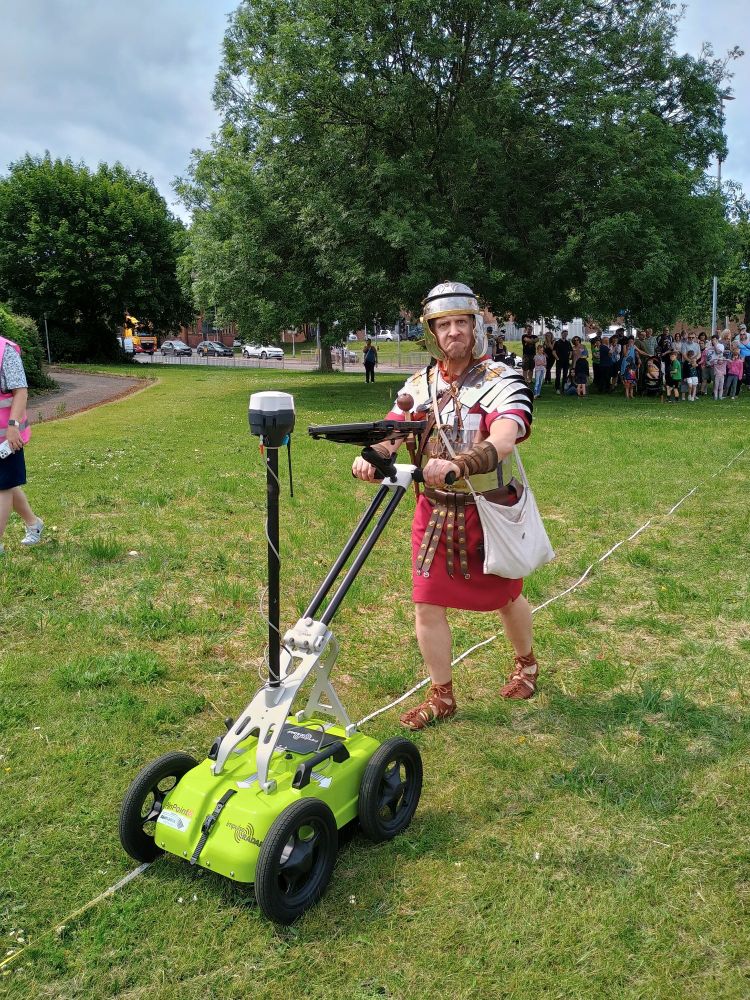 Roman Centurian pushing a Geophysics cart over a grassed area. He is pulling a funny face. A crowd is watching in the background. 