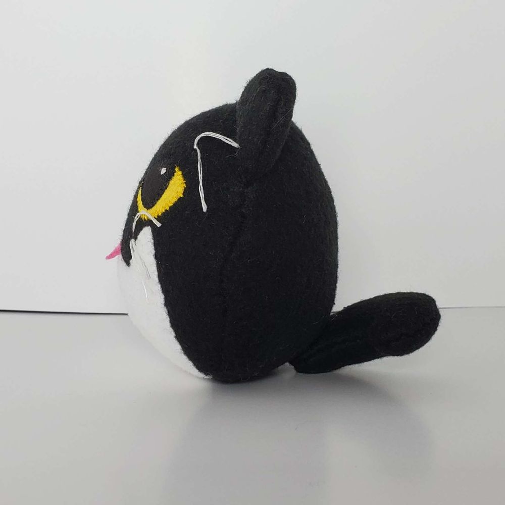 A surprisingly round plush of a black cat on a white background. The bunny ears have been removed. His eyes are yellow and his tummy is white. The plush is facing to the right and you can see his tail.