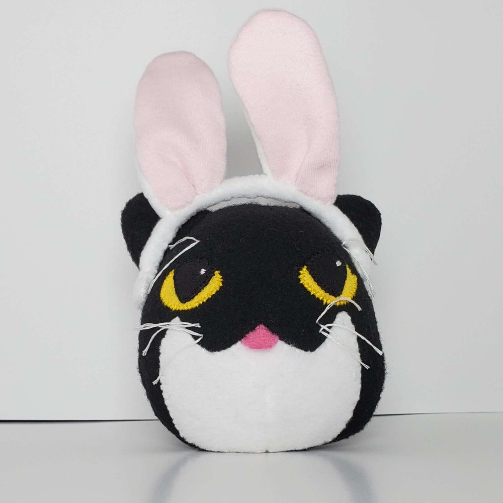 A surprisingly round plush of a black cat with a bunny ear headband on a white background. His eyes are yellow and his tummy is white. This is front facing and you can see his tongue sticking out.
