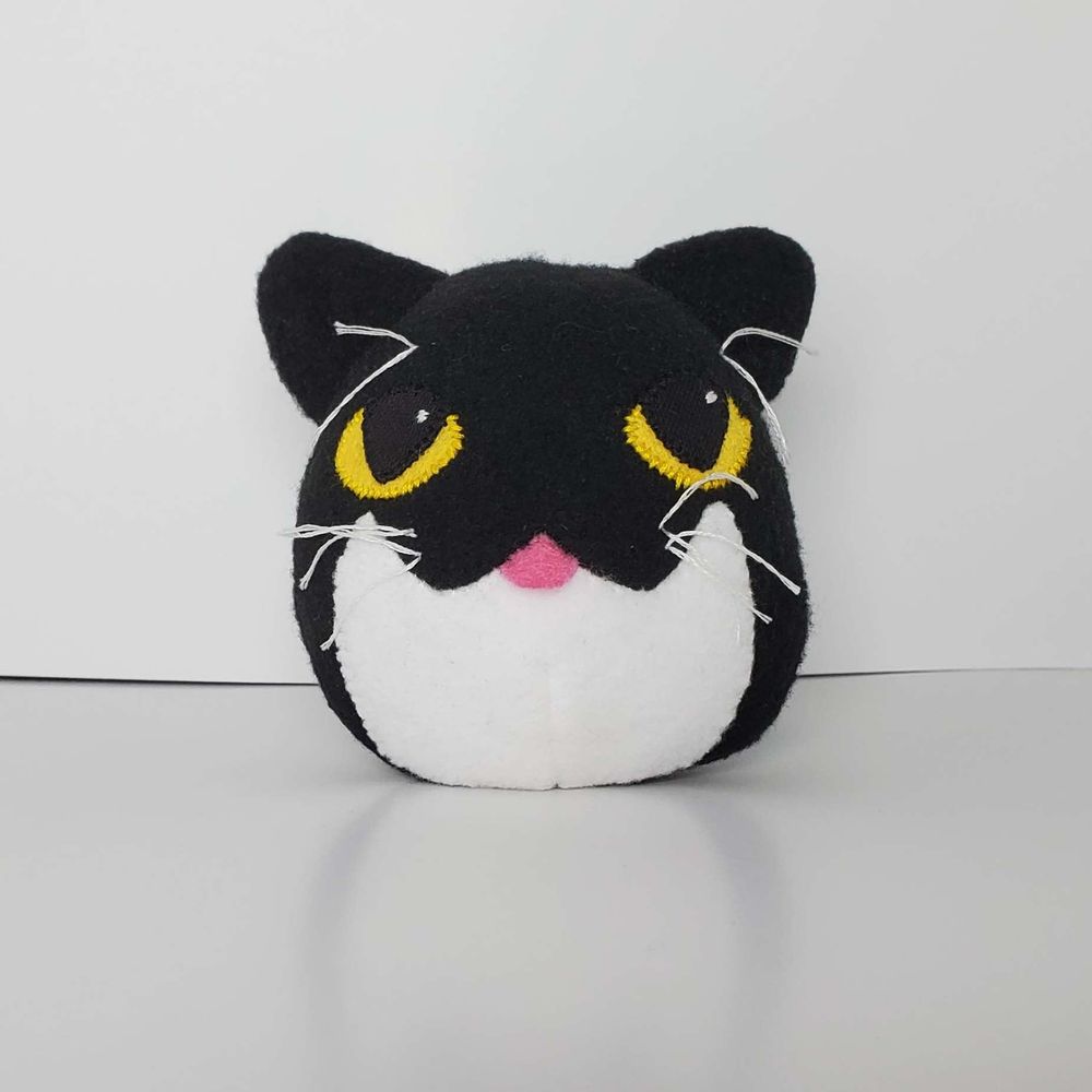 A surprisingly round plush of a black cat on a white background. The bunny ears have been removed. His eyes are yellow and his tummy is white. This is front facing and you can see his tongue sticking out.