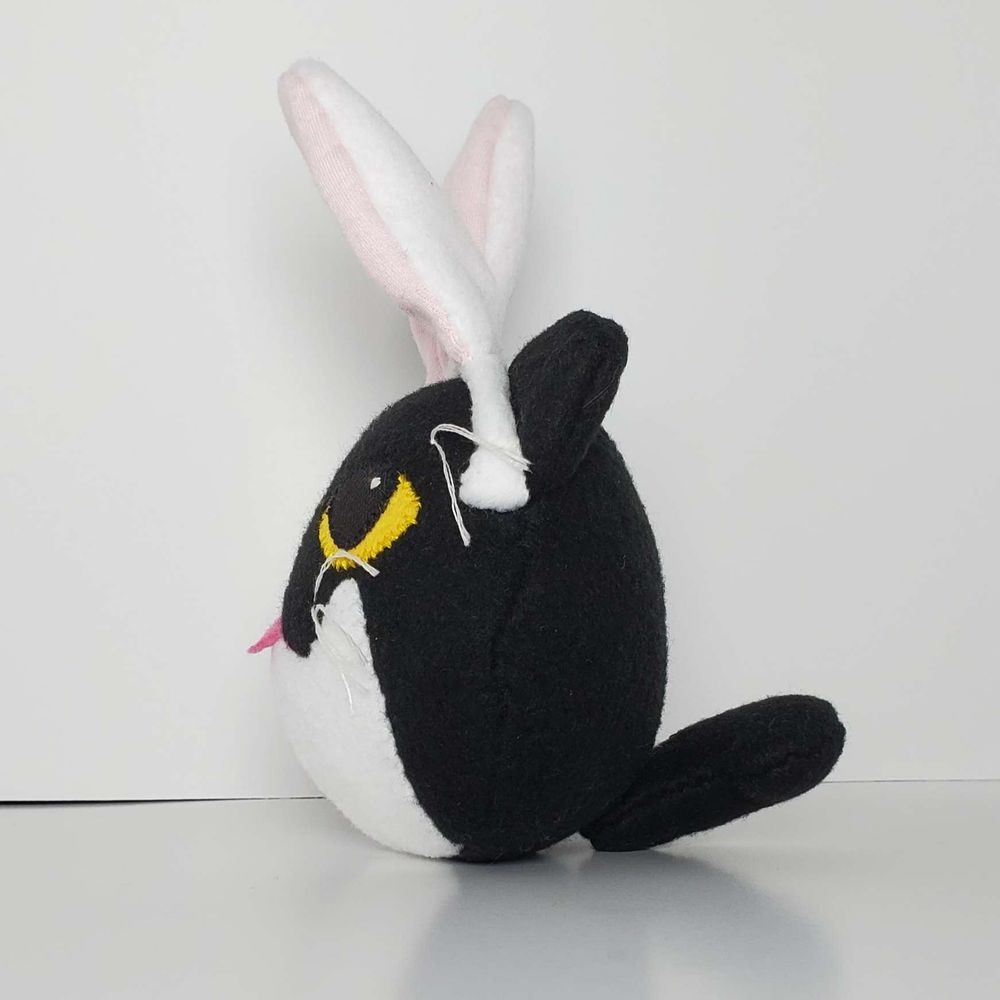 A surprisingly round plush of a black cat with a bunny ear headband on a white background. His eyes are yellow and his tummy is white. This is facing to the right, where you can see his tail. 