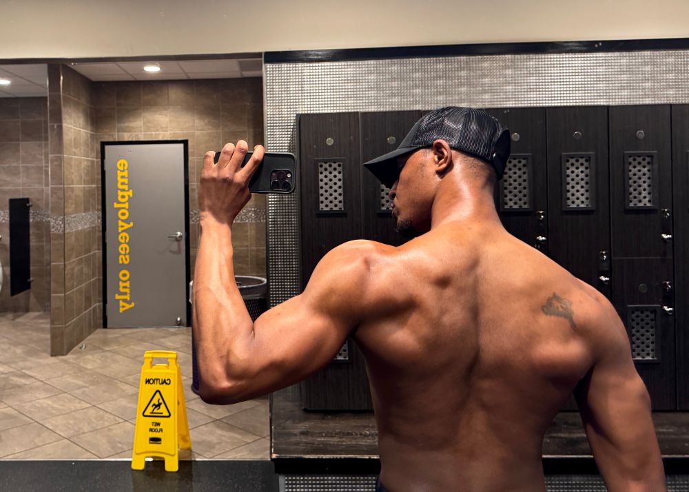 Antoine (the Designer Baddie, CFO of Shoulder Real Estate, Patron Saint of Not Giving a Fuck) in the gym locker room flexing shirtless with his cap pulled low. He’s slightly angled to the side, showing off wide back definition, full traps, and delts carved like architectural blueprints. The yellow “caution” sign in the mirror feels like a warning label for anyone unprepared for this much musculature.