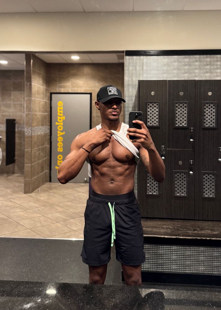 Antoine (CEO of Chest & Chaos Inc., that nigga, Mr. Fine Shyt) standing in front-facing form under locker room lights. He’s lifting his white tank top high to showcase tiddies in full spotlight, pecs round and commanding, abs ridged with precision. It’s time to weekend.