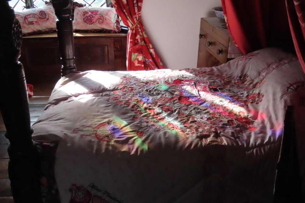 A colour photograph in landscape format. It shows the interior of a country house with a solid four-poster bed. On the bed is a white bedspread adorned with patchwork and embroidery created in 2022 for an exhibition. Coloured light from a nearby stained glass window fall on to the white linen.