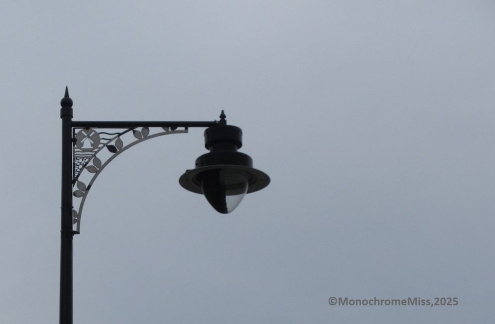 A colour photograph in landscape format. It looks monochrome, but is actually colour, but on a grey cloudy day without contrast. The view is up at a lampost with decorative metalwork. In the arch formed by the horizontal bar is a depiction of a windmill, with the sea behind and pebbles below. A row of pairs of leaves decorates the lower arch. Simple but a very nice way to recognise the building nearby.