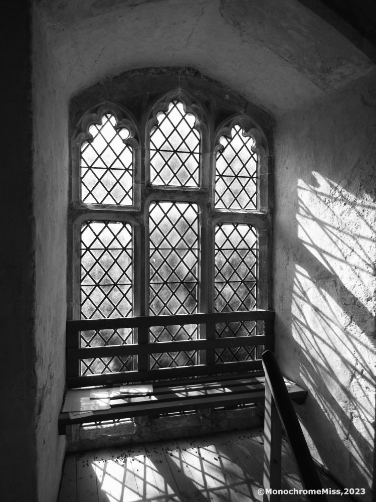 A black and white photograph in landscape format. It shows a leaded glass window in a tower. The light casts delicate shadows on the white wall.