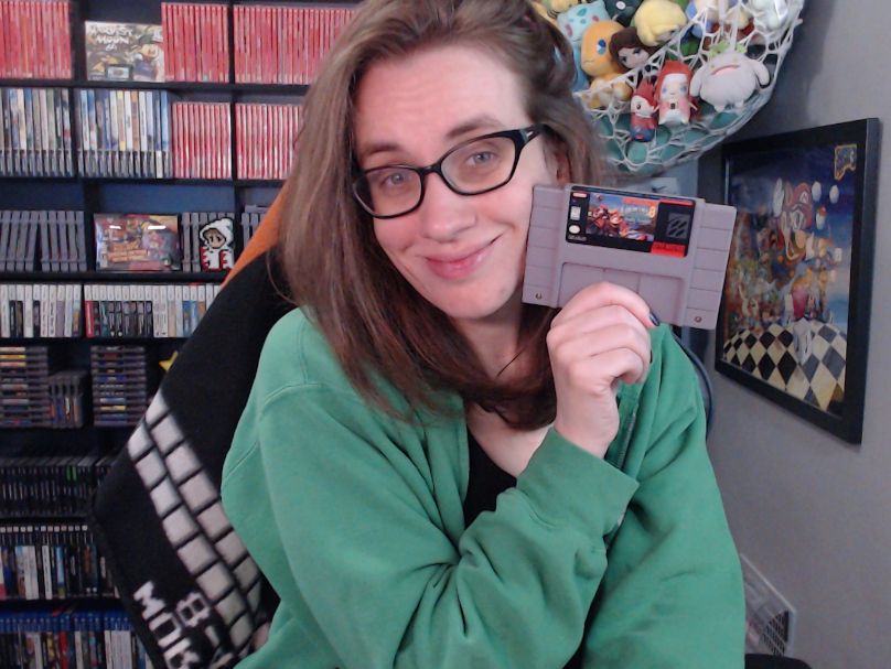 a picture of megan in her stream room. she is wearing a green hoodie and holding a copy of donkey kong country 3 for the super nintendo