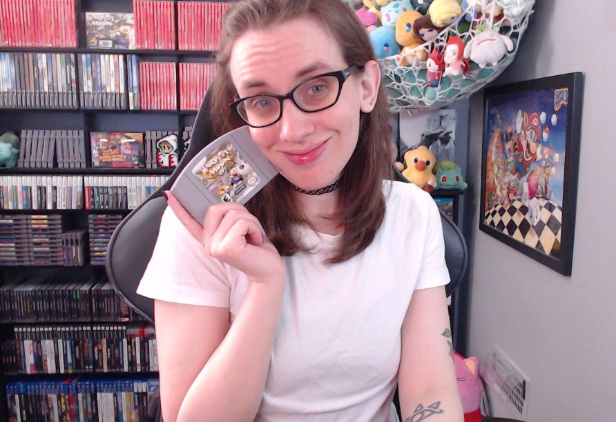 a photo of megan in her stream room! she is wearing a white tshirt and holding a copy of harvest moon 64 for the nintendo 64