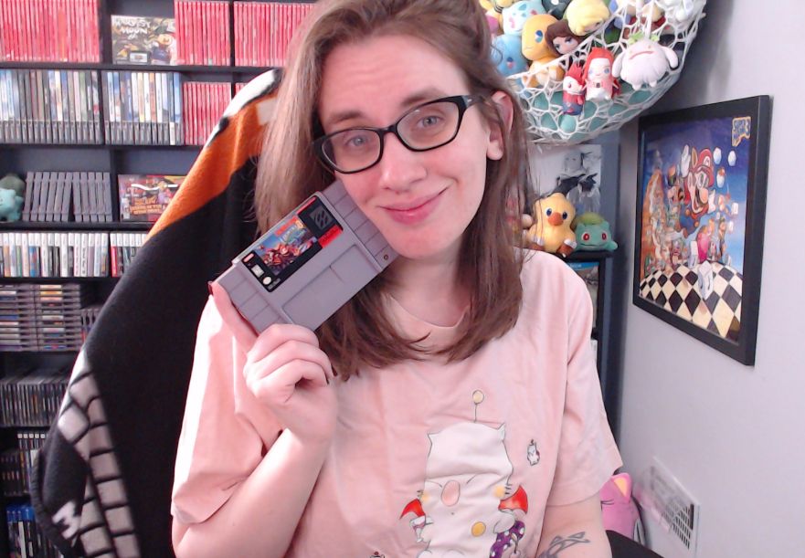 a picture of megan in her stream room. she is wearing a pink tshirt with artwork of the moogle from final fantasy vi on it and holding a copy of donkey kong country 3 for the super nintendo