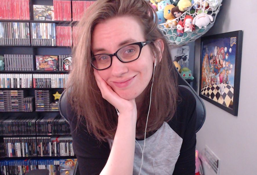 a picture of megan in her stream room. she is wearing black thick-framed glasses and a black and grey baseball style halfsleeve teeshirt.