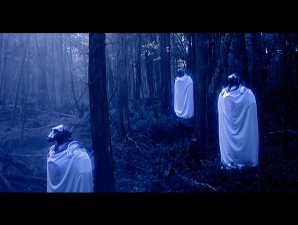 Three animal-headed statues, shrouded in white cloaks, in a blue tinted forest