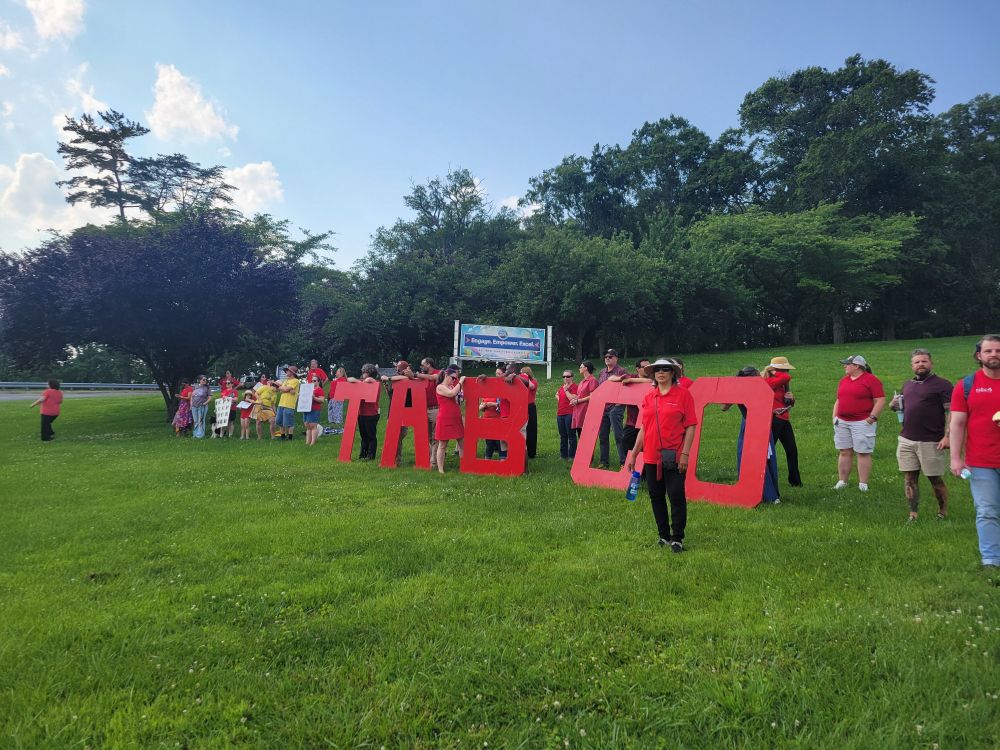 The lawn in front of the Board of Education's campus. There are about 25 protestors scattered on the hill behind 5 large, red, wooden letters that spell out TABCO. 