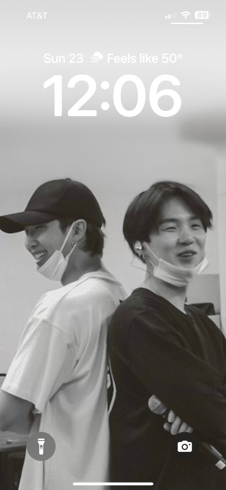 iPhone lockscreen showing Namjoon and yoongi standing back to back smiling. Namjoon is on the left wearing a white t shirt a black cap and a face mask pulled below his chin. Yoongi is wearing a black long sleeve and also has a face mask pulled below his chin